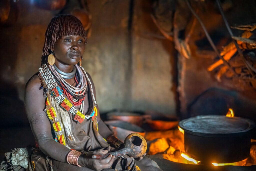 Hüttenbewohnerin in traditioneller Kleidung in Afrika, mit Kochgeschirr am Feuer.