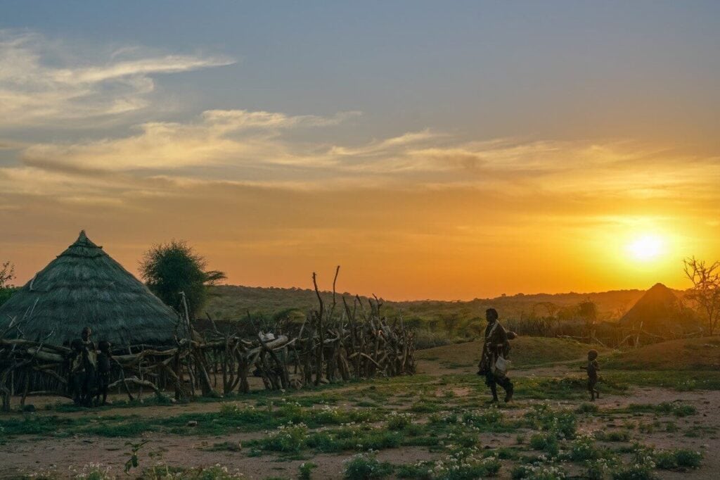 Sonnenuntergang über einem afrikanischen Dorf mit traditionellen Hütten und Dorfbewohnern.