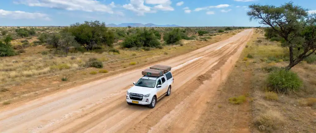 Ein weißer Geländewagen fährt auf einer staubigen Autobahn durch die afrikanische Savanne, umgeben von trockenen Büschen und Akazienbäumen.