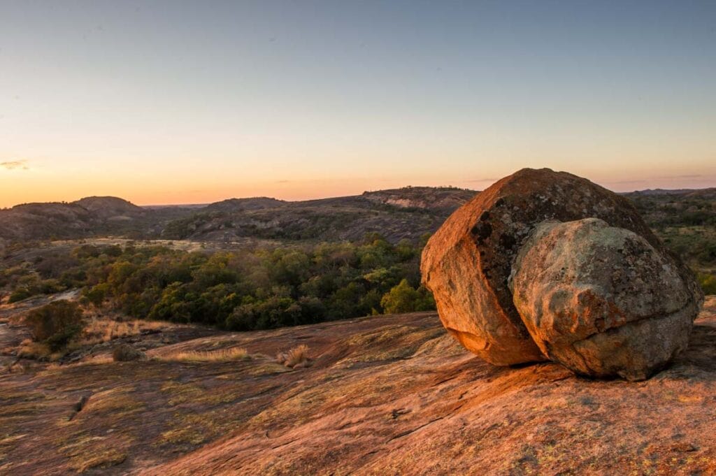 Sonnenuntergang über afrikanischer Wildnis mit rot-orangenen Felsen und Buschland.