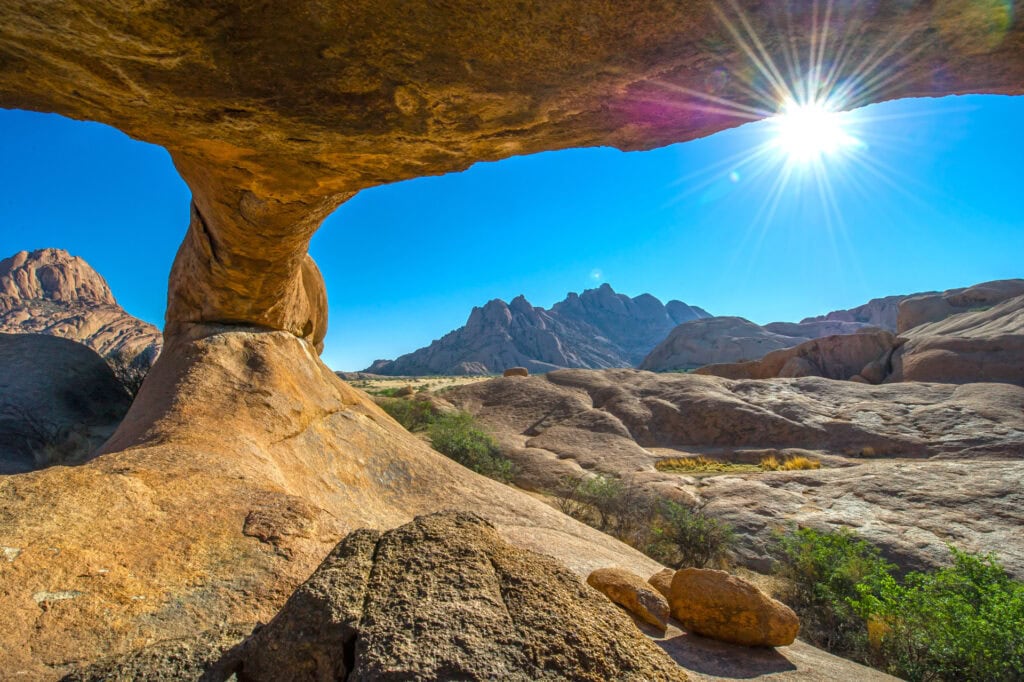 Oase in der Wüste mit beeindruckender Felsformation und Bergen, Sonnenlicht in der Wüstenlandschaft.