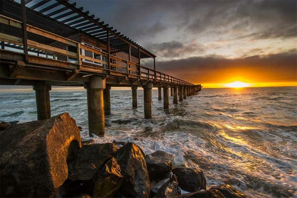 Sunset am afrikanischen Strand mit Pier, Wellen und Felsen.