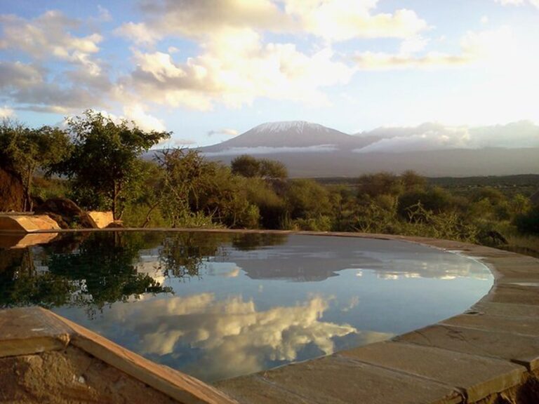 Panoramaaufnahme eines Swimmingpools vor einem afrikanischen Busch, im Hintergrund der Mount Kilimanjaro bei Sonnenaufgang.