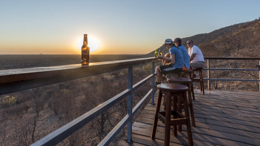 Menschen genießen Sonnenuntergang auf einer Safari-Deck in Afrika, Blick auf die Wildnis, Tierbeobachtung, Naturerlebnis.