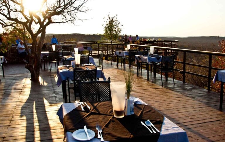 Altar Text: Elegantes Restaurant auf einer Dachterrasse mit Blick auf die afrikanische Wildnis bei Sonnenuntergang.