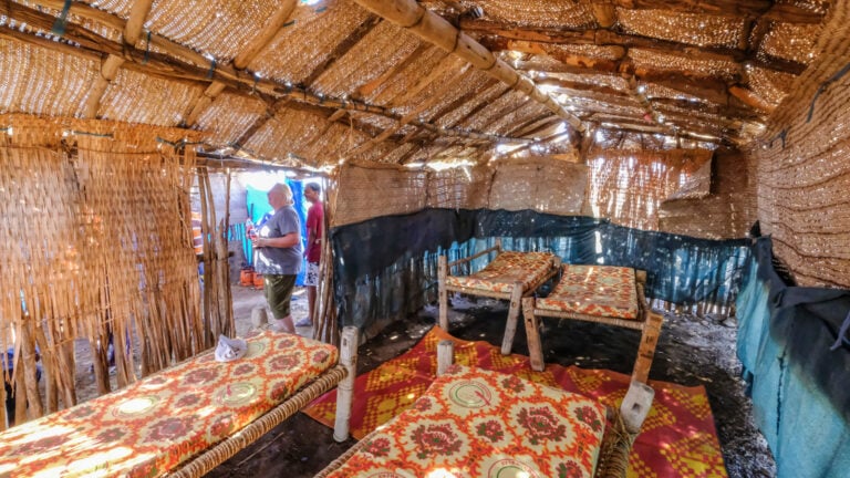 ALT Text: Traditionelles afrikanisches Zelt mit Holzliegen, Bastwänden und Sonnenlicht.