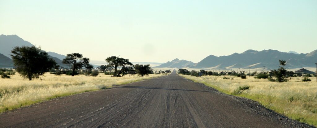Lange Straße durch Afrikas Wüstenland, umgeben von Gras und Akazien, im Hintergrund Berge.