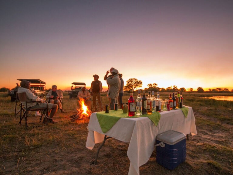 Alt: Safari-Gruppe bei Sonnenuntergang mit Camping, Lagerfeuer und Safari-Fahrzeugen in Afrika.
