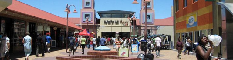 Die Menschen gehen durch ein Einkaufszentrum mit bunten Gebäuden, einem zentralen Brunnen und einem Schild mit der Aufschrift Wohlwillplatz über dem Haupteingang. Der Himmel ist klar und blau.