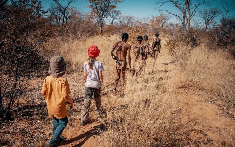 Alt: Gruppe von Touristen und Einheimischen auf Safari in Afrika, umgeben von Trockenstrauch und Bäumen.