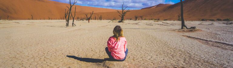 ALT: Mädchen in roter Jacke sitzt in der namibischen Wüste, umgeben von vertrockneten Bäumen.