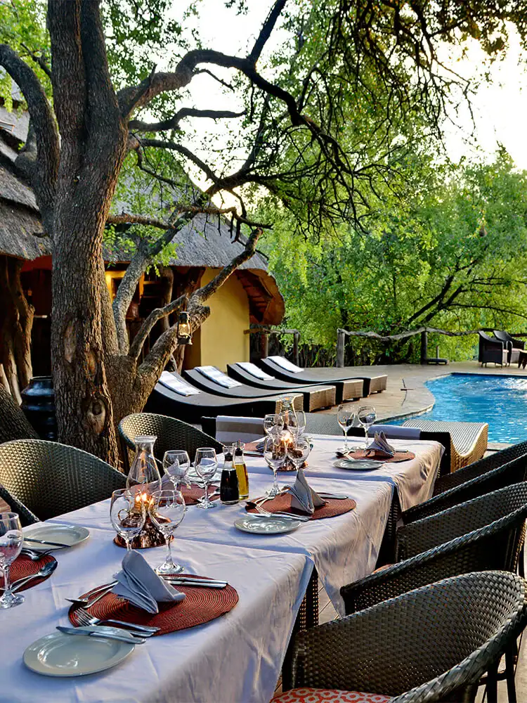 Blick auf ein afrikanisches Lodge-Restaurant mit Tischgedeck und Pool, umgeben von Natur.