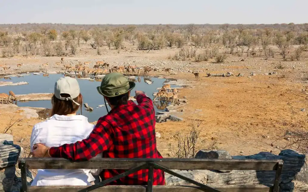 Zwei Touristen beobachten Wildtiere an einem Wasserloch in Namibia, umgeben von trockener Savanne.