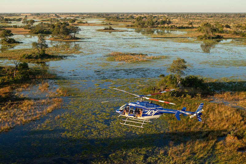 Helikopter fliegt über afrikanische Landschaft mit Wasser und Bäumen.