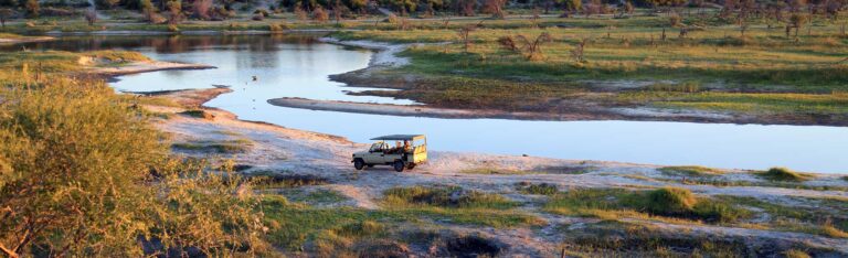 Ein Safarifahrzeug, das am sandigen Ufer eines Flusses in der grasbewachsenen Landschaft des Makgadikgadi-Pans-Nationalparks geparkt ist.