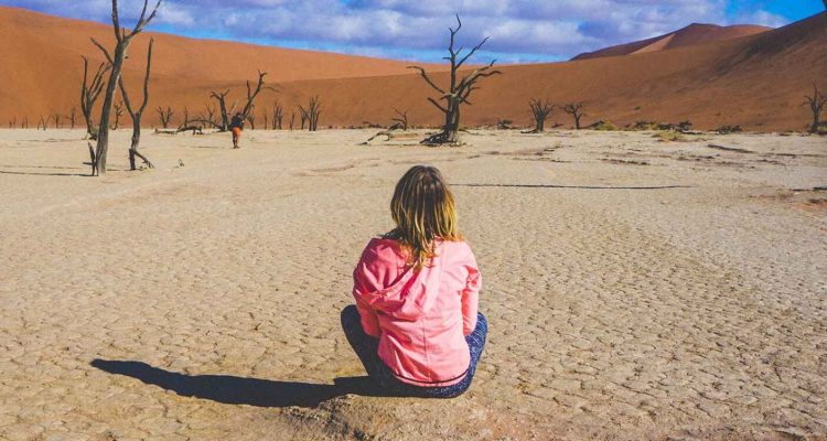 ALT: Mädchen in roter Jacke sitzt in der namibischen Wüste, umgeben von vertrockneten Bäumen.